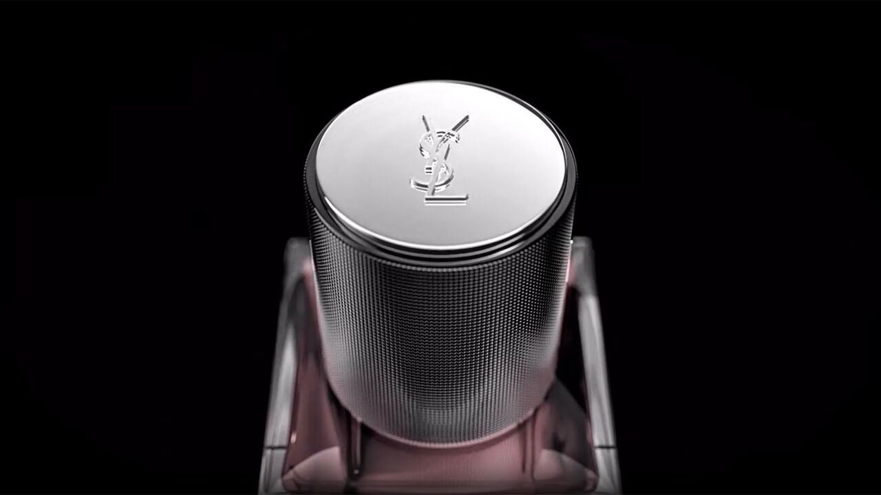 A close-up view of a silver, textured perfume cap with the YSL logo prominently embossed on top, set against a dark background.
