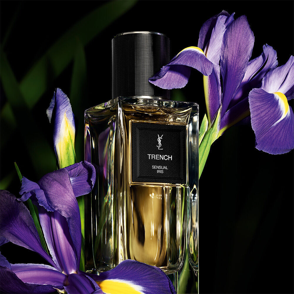 A golden-toned perfume bottle labeled "TRENCH SENSUAL IRIS" by YSL, prominently displayed alongside vibrant purple irises.
