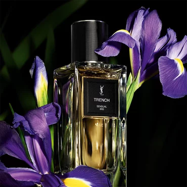 A golden-toned perfume bottle labeled "TRENCH SENSUAL IRIS" by YSL, prominently displayed alongside vibrant purple irises.