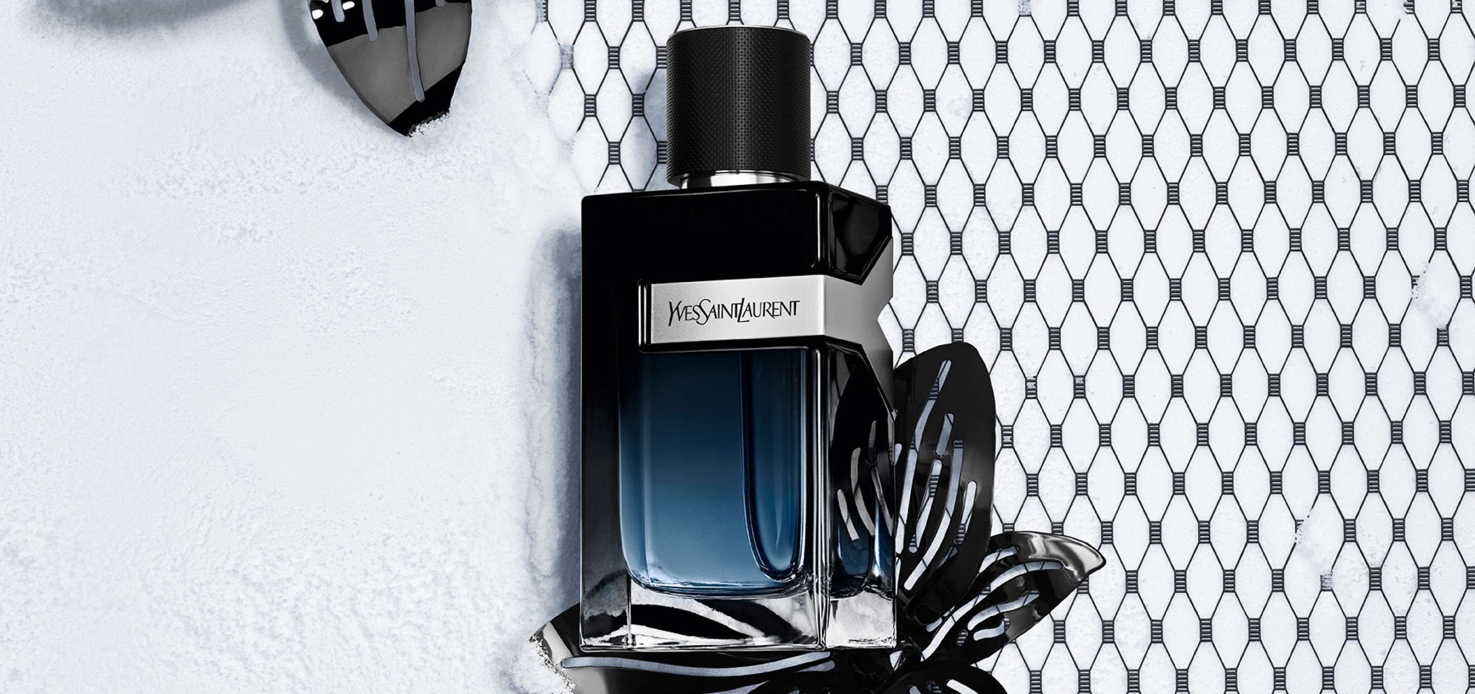 A stylish bottle of Yves Saint Laurent perfume sits on a textured surface, surrounded by black decorative elements and a mesh background. The bottle features a sleek design with a dark cap and a gradient blue to clear glass.