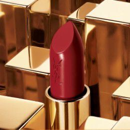 A close-up of a vibrant red lipstick with a sleek, gold casing, set against a backdrop of shiny gold blocks. The lipstick features the YSL logo embossed on its side.
