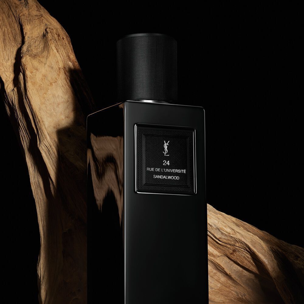 A sleek, rectangular black perfume bottle labeled "24 RUE DE L'UNIVERSITE SANDALWOOD" by YSL, positioned against a backdrop of natural, rustic wood.