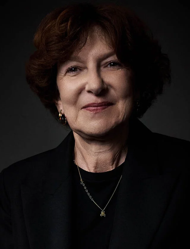 A smiling woman with short brown hair, wearing a dark suit and a delicate necklace, is seated on a dark leather chair.