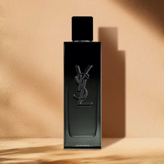 A sleek, rectangular black perfume bottle with a prominent embossed black YSL logo on the front. The words "EAU DE PARFUM" are visible near the bottom. The bottle has a black cap and is standing on a light wooden surface against a warm, plain beige background, with shadows cast to the right.