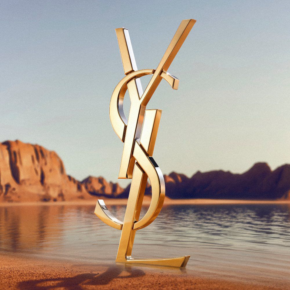 A large, iconic 3D gold YSL (Yves Saint Laurent) logo standing on a desert shoreline. The polished gold monogram reflects the sunlight and is positioned in front of a calm body of water with dramatic red rock formations and desert mountains in the background under a clear sky.