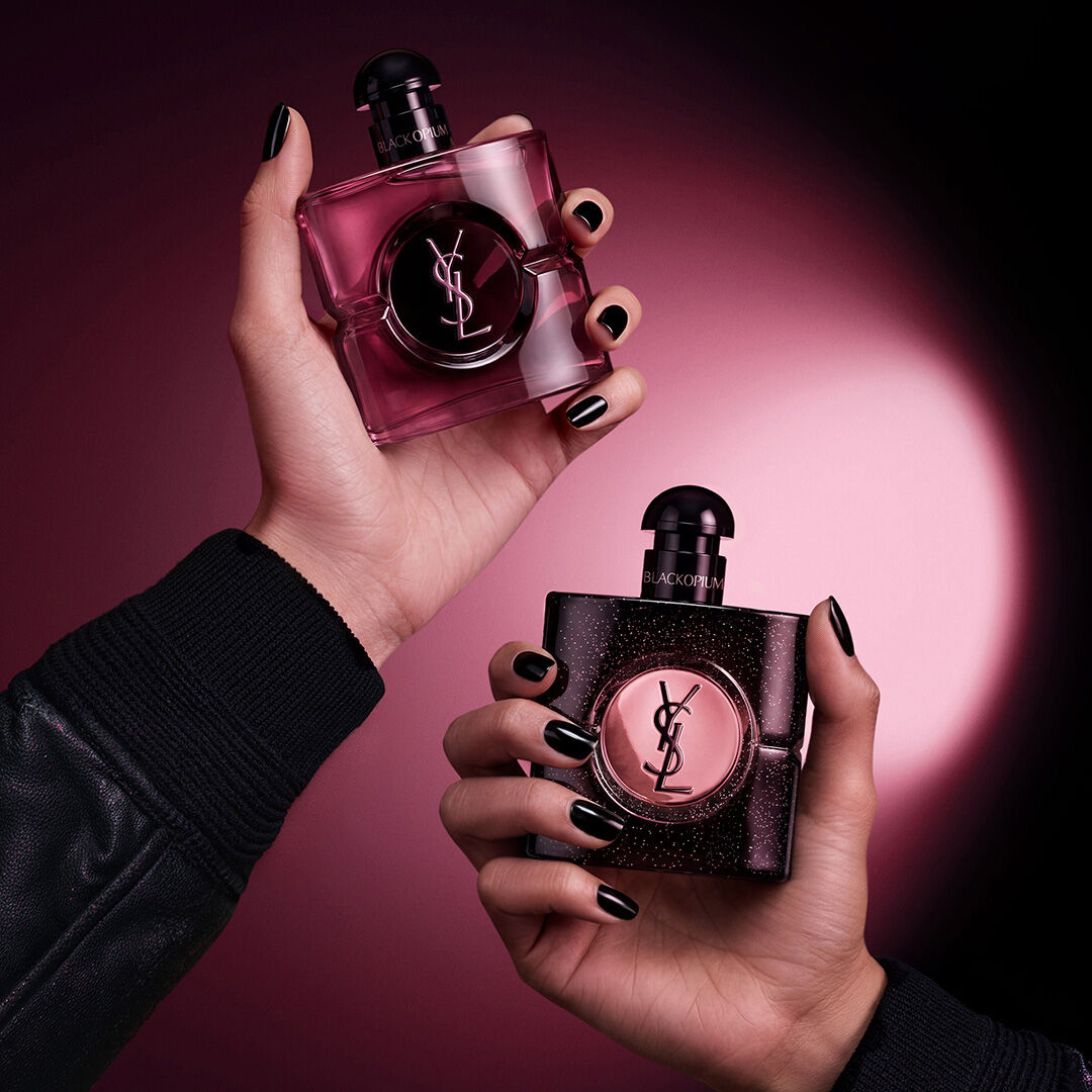Two hands holding Yves Saint Laurent Black Opium fragrance bottles against a dark pink and black background.