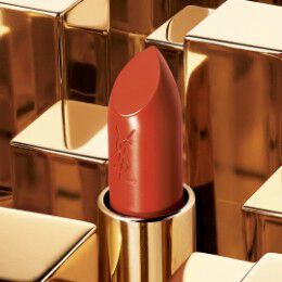 A close-up of a red lipstick in a gold tube, set against a background of shiny gold geometric shapes. The lipstick features the YSL logo etched on its side.