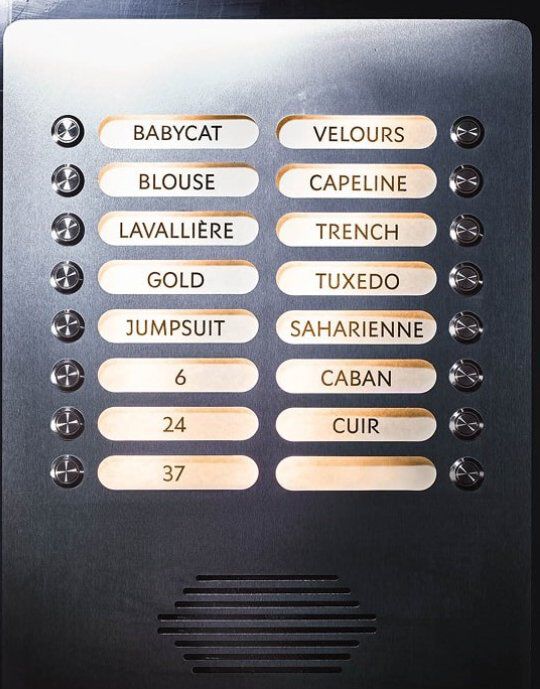 Metallic panel display with illuminated buttons, each labeled with a word referencing YSL fashion. Labels include 'BABYCAT', 'TUXEDO', 'SAHARIENNE', and 'MUSE', among others. The panel features small circular buttons and a speaker grille at the bottom, resembling an elegant intercom system.
