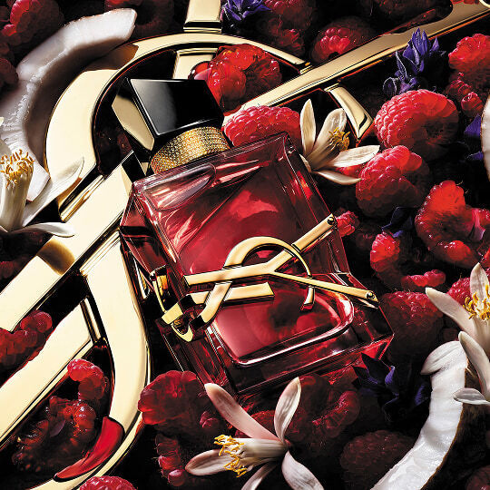 The pink Yves Saint Laurent Libre Berry Crush bottle surrounded by raspberries, white orange blossoms, and coconut slices on a gold logo background.