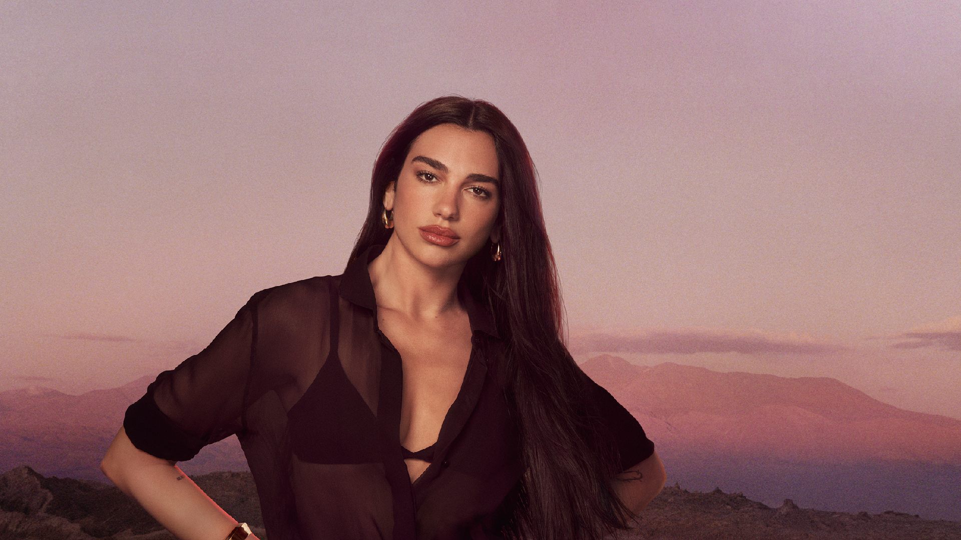 Dua Lipa posing in a black sheer shirt against a sunset mountain landscape.