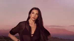 Dua Lipa posing in a black sheer shirt against a sunset mountain landscape.