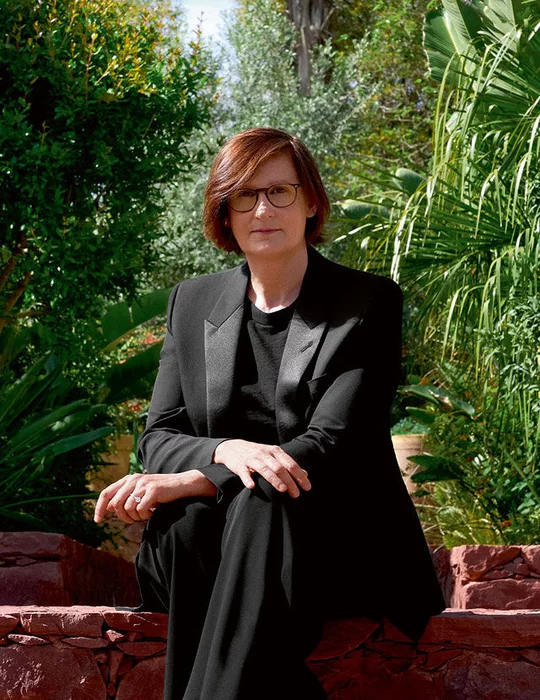 A woman with reddish-brown hair and glasses sits on a stone wall, wearing a black suit. She is looking at the viewer, with a lush green garden in the background.
