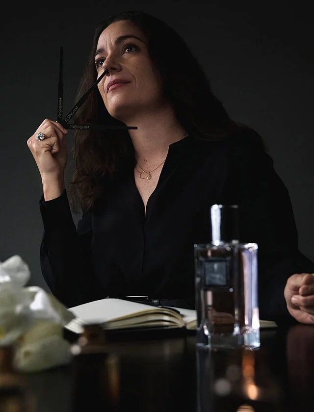 A woman in a black shirt holds a perfume blotter, looking up thoughtfully, with a notebook, perfume bottle, and flowers on the table.