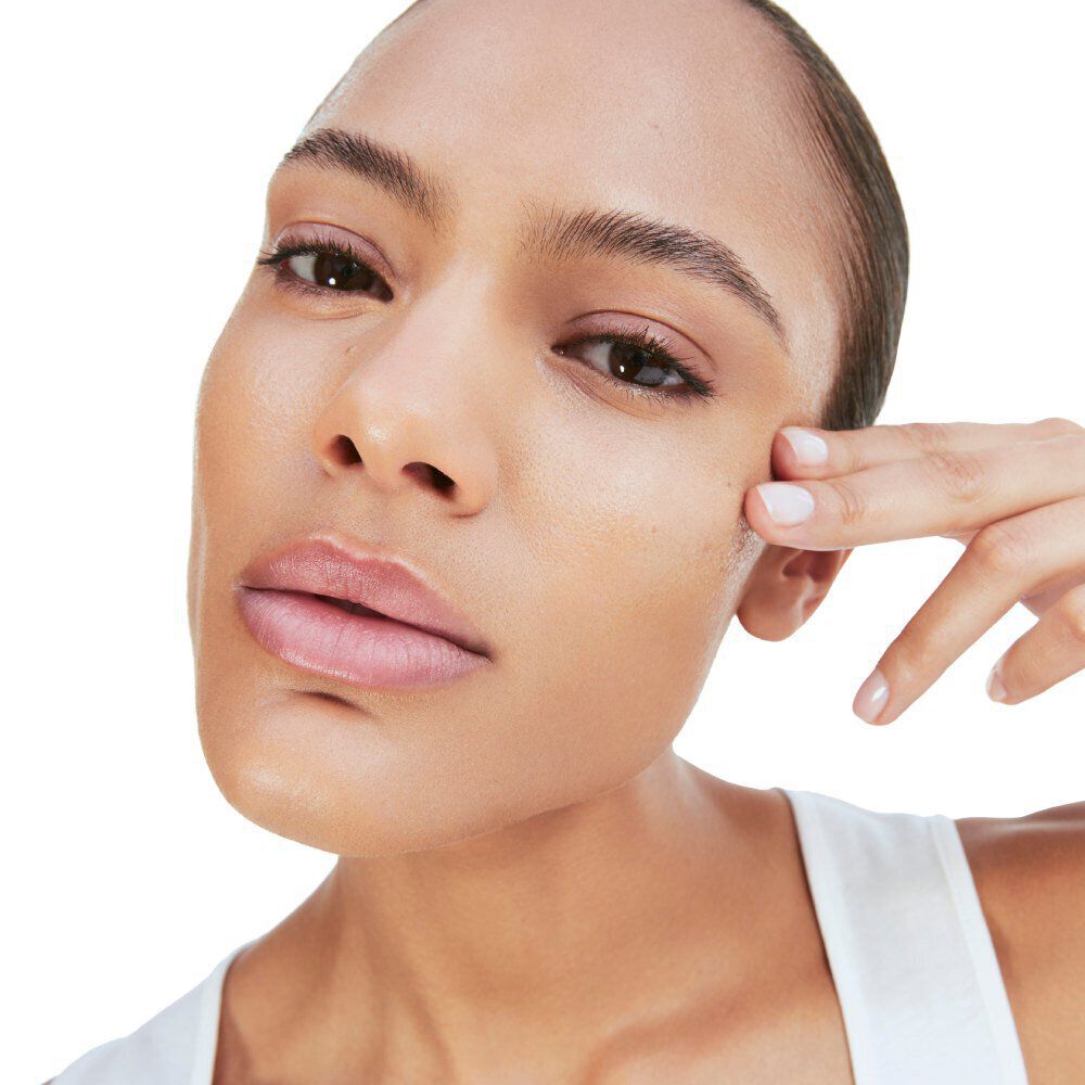 A close-up of a person with a smooth complexion, gently touching their cheek with one hand, looking directly at the camera. They have a neutral expression and are wearing minimal makeup.