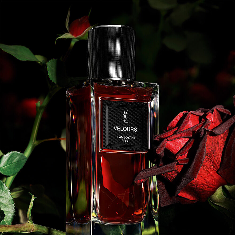 A dark red perfume bottle labeled "VELOURS FLAMBOYANT ROSE" by YSL, with a vibrant red rose in the blurred background.