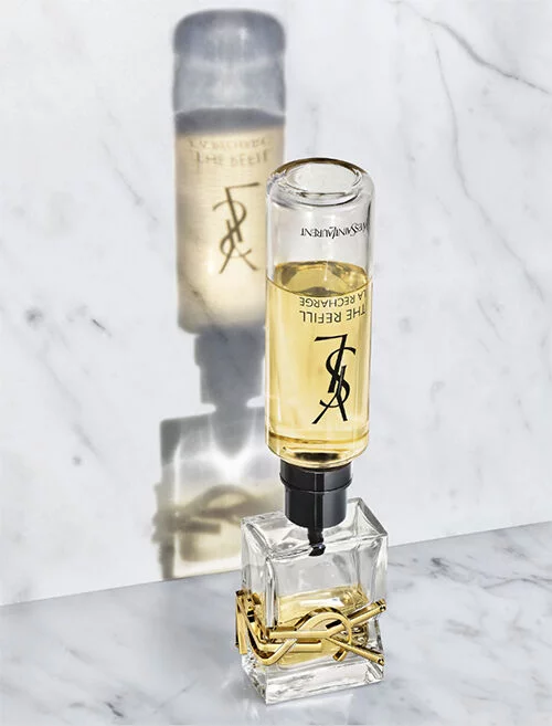 A glass perfume bottle with a silver cap sits on a marble surface, casting a shadow. The bottle is partially filled with a golden liquid and features the Yves Saint Laurent logo.