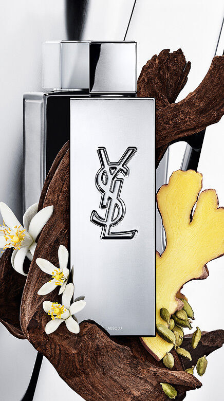 YSL Y L'ABSOLU perfume bottle surrounded by dark wood, white orange blossom flowers, ginger, and cardamom pods.