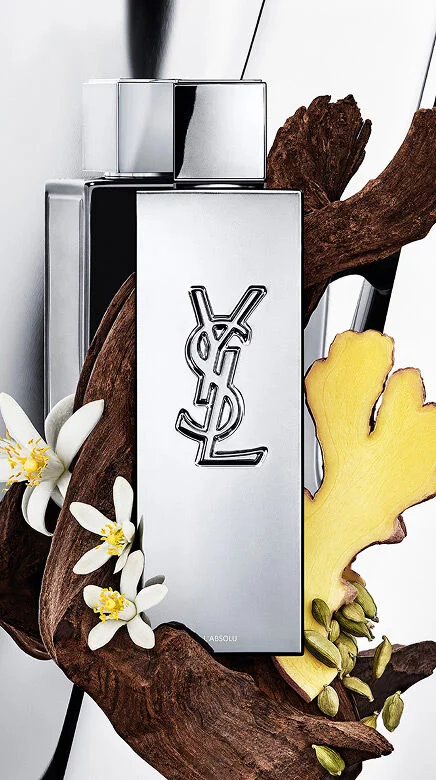 YSL Y L'ABSOLU perfume bottle surrounded by dark wood, white orange blossom flowers, ginger, and cardamom pods.