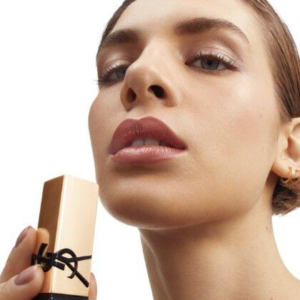 A close-up of a woman showcasing a beauty product, holding a gold-capped makeup container near her face while wearing a subtle makeup look.