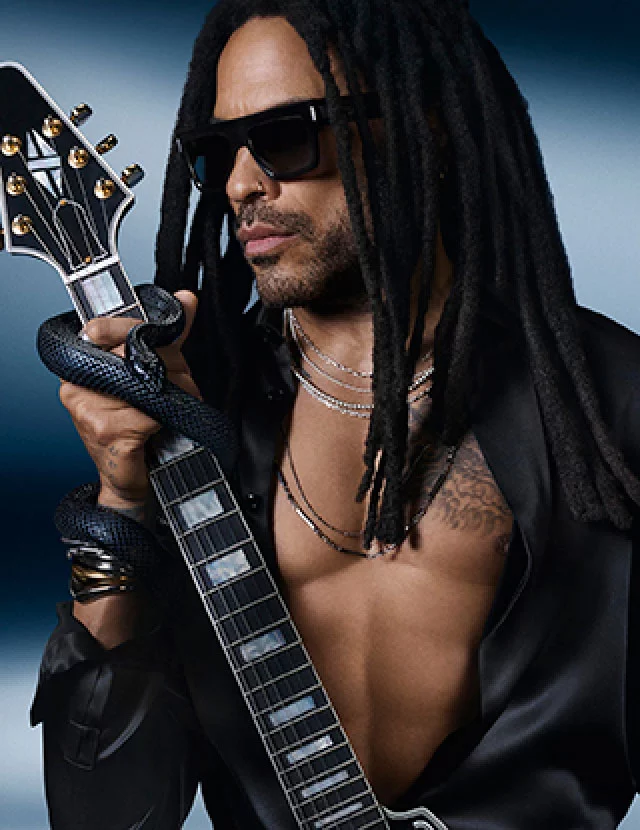 A man with long dreadlocks wearing sunglasses holds an electric guitar, with a snake wrapped around his arm. He is dressed in a black shirt and appears against a blurred blue background.