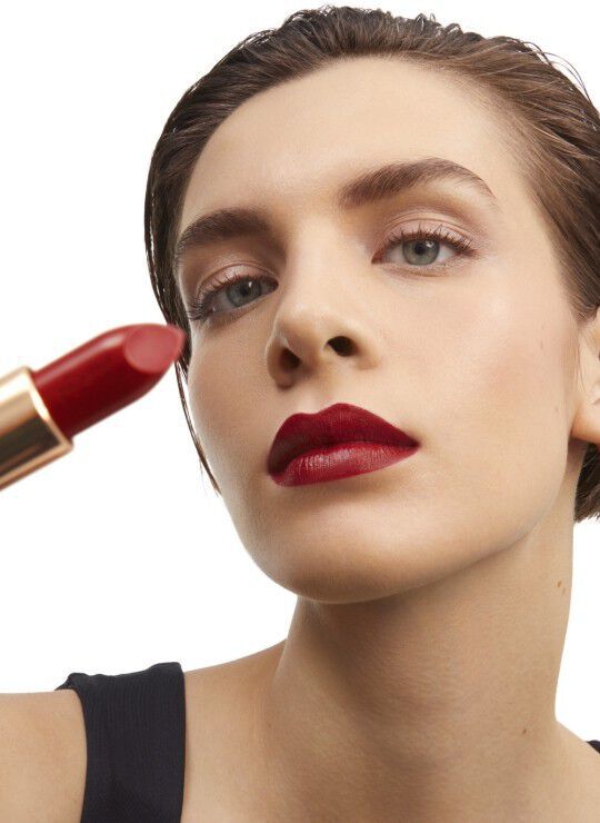 A close-up portrait of a model applying red lipstick, showcasing her bold lip color and flawless skin, with a focus on the lipstick tube she is holding.