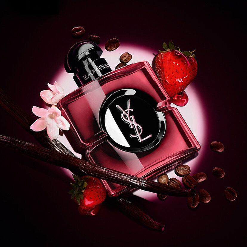 YSL Black Opium perfume bottle surrounded by coffee beans, strawberries, vanilla pods, and white flowers on a dark background.