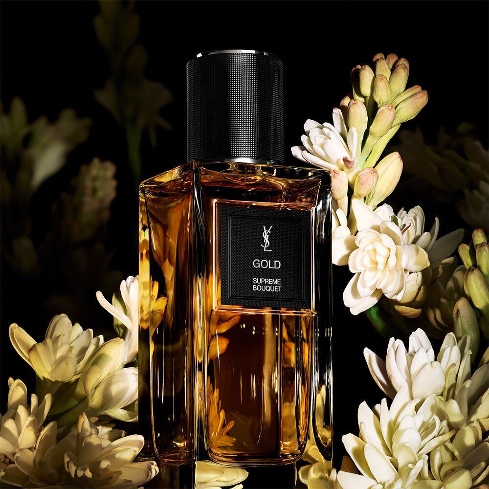A golden-toned perfume bottle labeled "GOLD SUPREME BOUQUET" by YSL, richly surrounded by a cluster of white tuberose flowers.