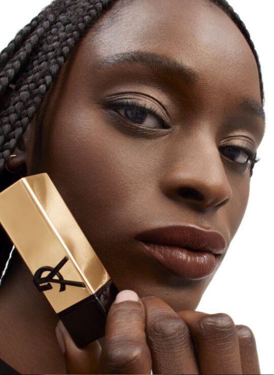 A close-up portrait of a woman with braided hair, showcasing a makeup product in her hand. She has a neutral makeup look, emphasizing her lips and eyes, with a soft-focus background. The product has a gold and black packaging, featuring the Yves Saint Laurent logo.