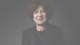 A close-up portrait of a smiling woman with short brown hair, wearing a dark suit and a delicate necklace, against a dark background.