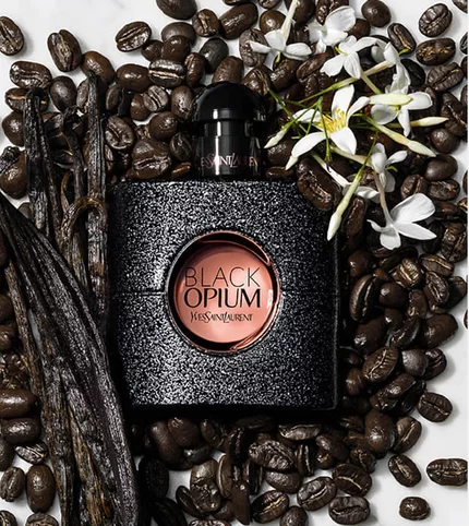 A bottle of Yves Saint Laurent Black Opium perfume surrounded by coffee beans and vanilla pods, with a white flower accenting the scene.