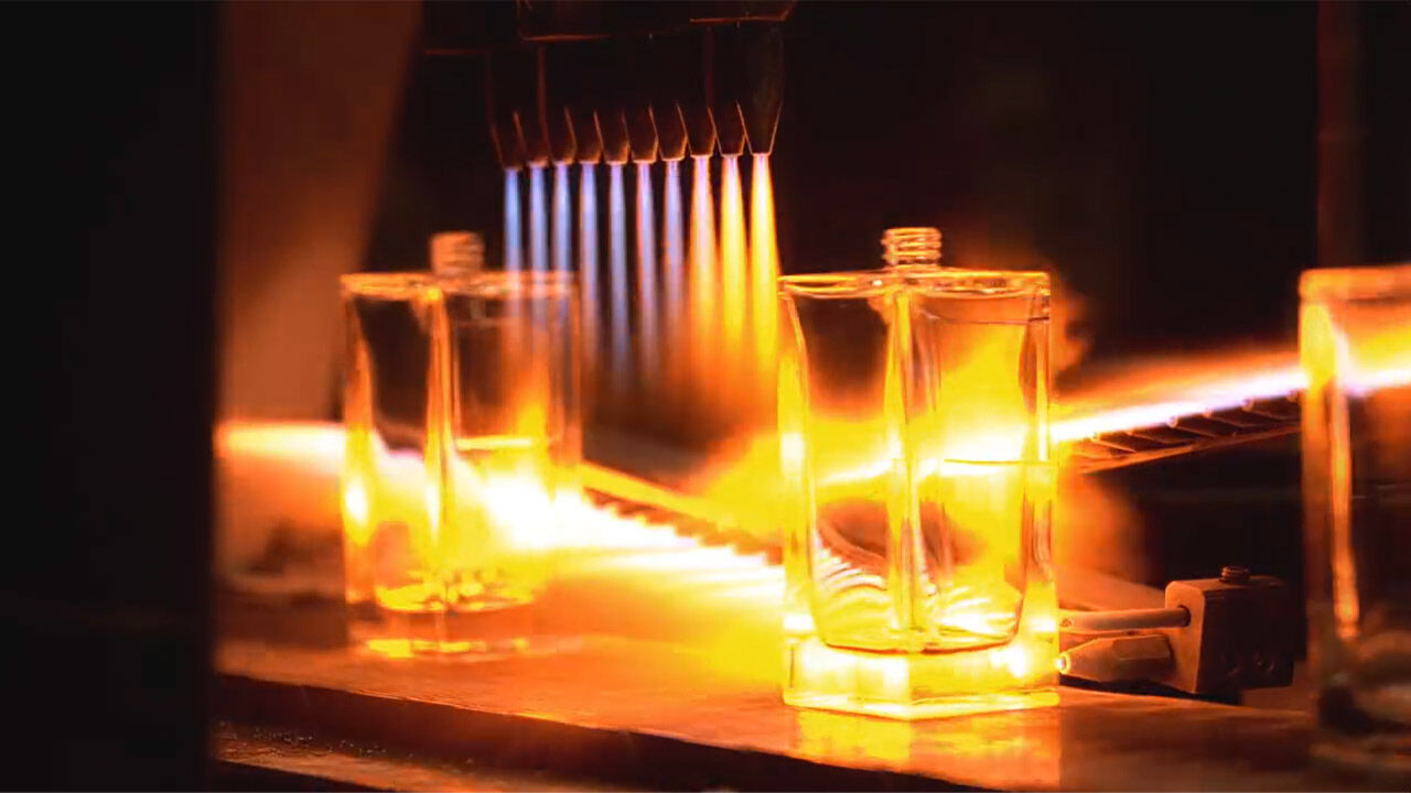 A close-up of a clear rectangular perfume bottle being intensively heated by flames from several nozzles, highlighting the industrial process with an orange glow.