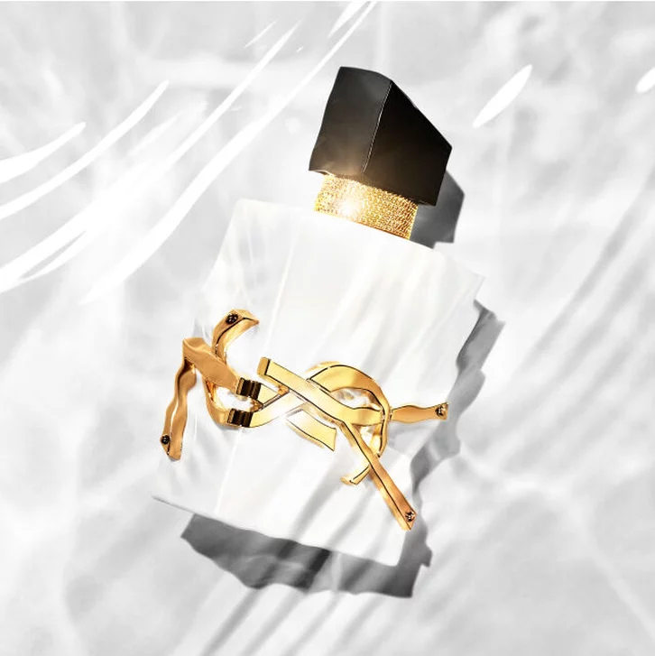 A stylish perfume bottle with a sleek white design, adorned with a gold accent, lying on a light background with water-like reflections.