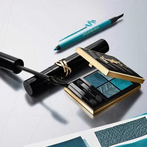 A luxury YSL beauty flat lay featuring an open gold-trimmed eyeshadow palette with blue and teal shades, a black mascara wand with product, and a teal eyeliner pencil, all arranged on a reflective silver surface.