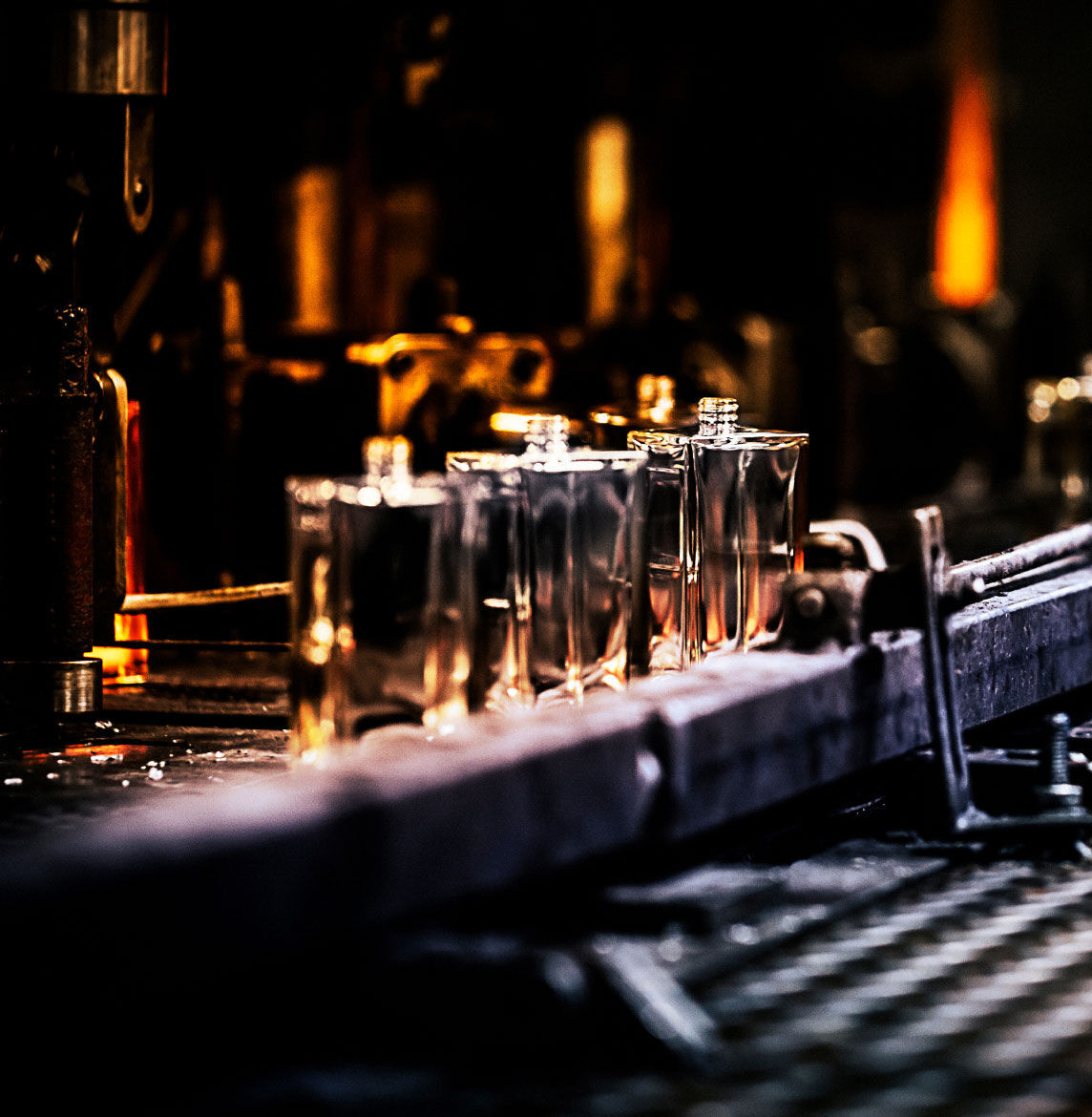 Multiple clear perfume bottles, glowing with an amber hue from behind, are lined up on a conveyor belt or processing line in a dimly lit factory.