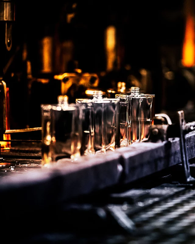 Multiple clear perfume bottles, glowing with an amber hue from behind, are lined up on a conveyor belt or processing line in a dimly lit factory.
