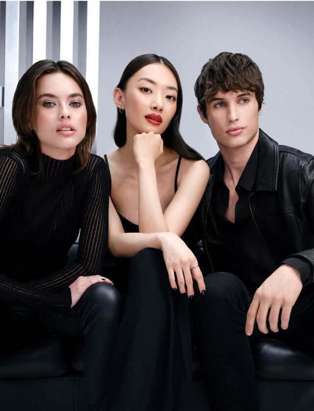 Three stylish young models in black outfits sitting on a black leather sofa against a grey studio background.