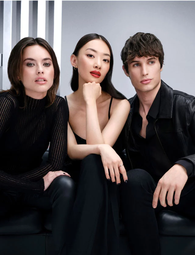 Three stylish young models in black outfits sitting on a black leather sofa against a grey studio background.