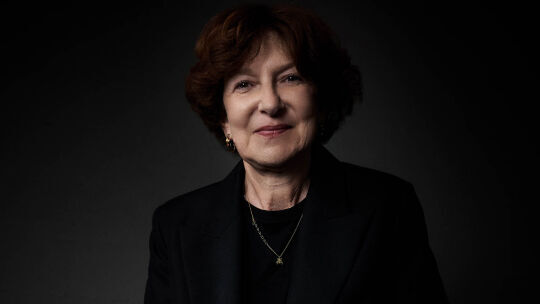 A close-up portrait of a smiling woman with short brown hair, wearing a dark suit and a delicate necklace, against a dark background.
