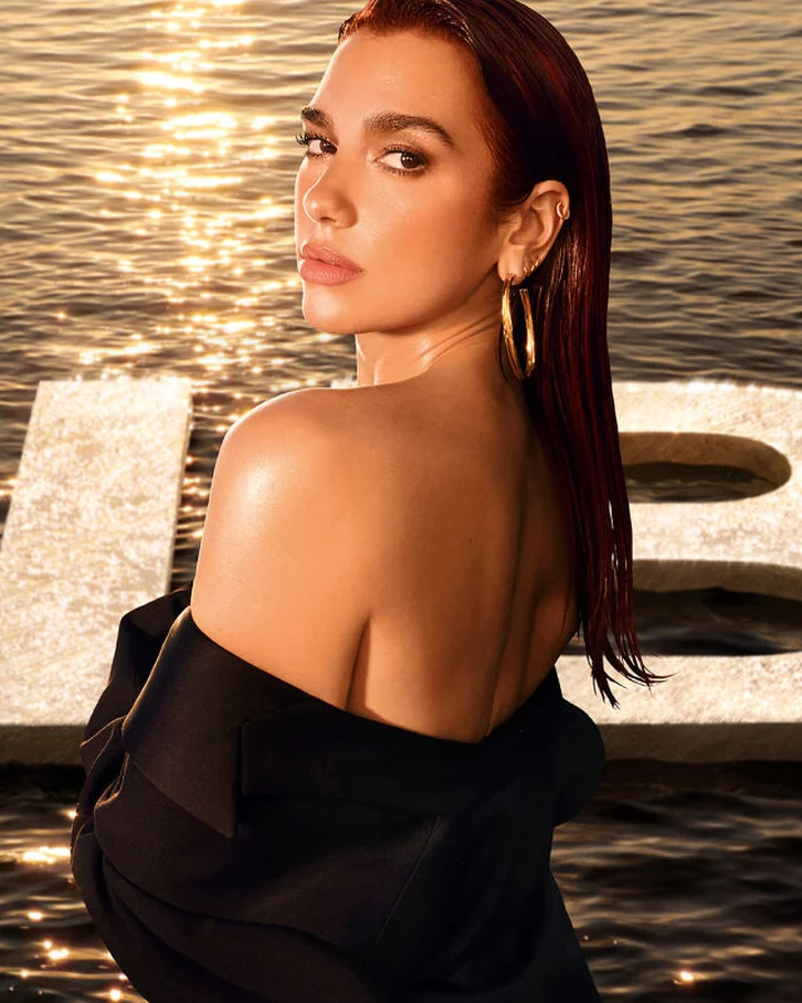 A woman with long, dark hair gazes back over her shoulder while standing in front of large letters spelling "LIBRE," with a shimmering water background reflecting warm light.