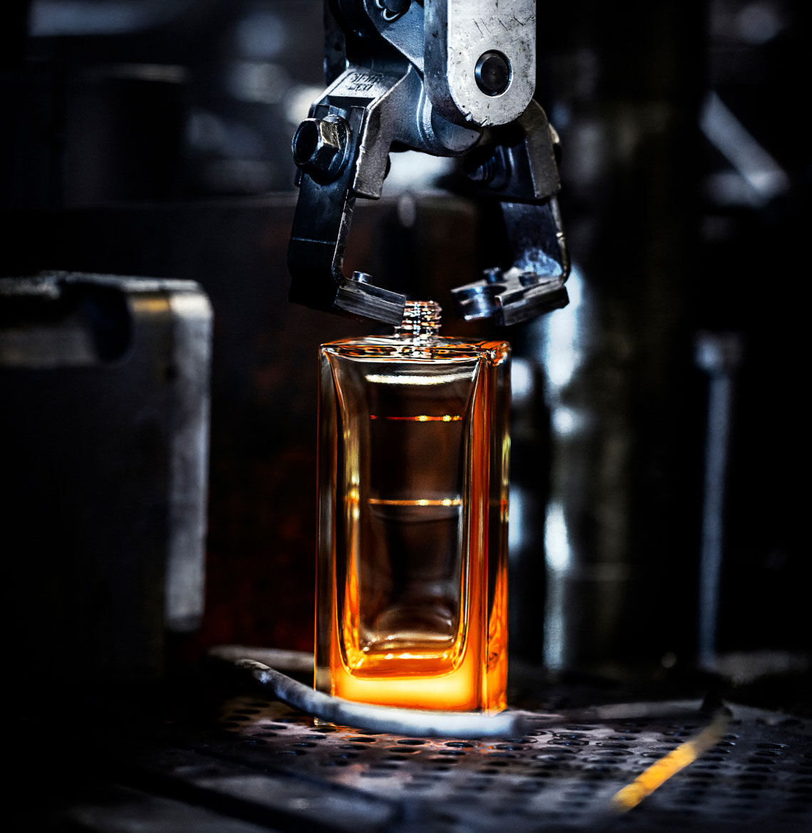 A robotic arm lowers a textured cap onto a glowing amber perfume bottle in a manufacturing setting, emphasizing precision in production.
