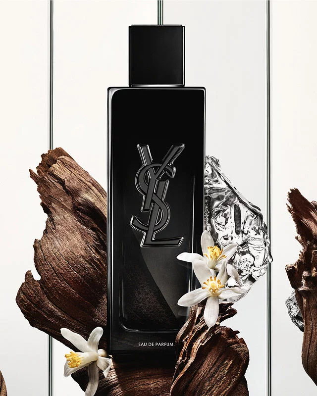 A black Yves Saint Laurent Eau de Parfum bottle with the YSL logo, surrounded by dark wood pieces, white delicate flowers, and a crumpled silver reflective object.
