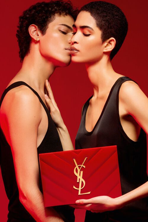 A man and a woman in sleek black outfits lean in for a kiss, holding a red YSL makeup case against a deep red background.