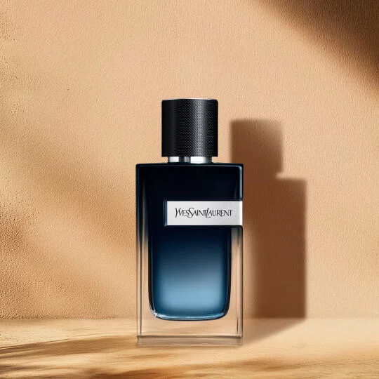 A rectangular perfume bottle with a dark blue liquid that transitions to a lighter blue towards the bottom. It features a textured black cap, a silver band around the neck, and a silver label on the front that reads "YVES SAINT LAURENT". The bottle rests on a light wooden surface, with soft shadows cast behind it, against a warm beige background.
