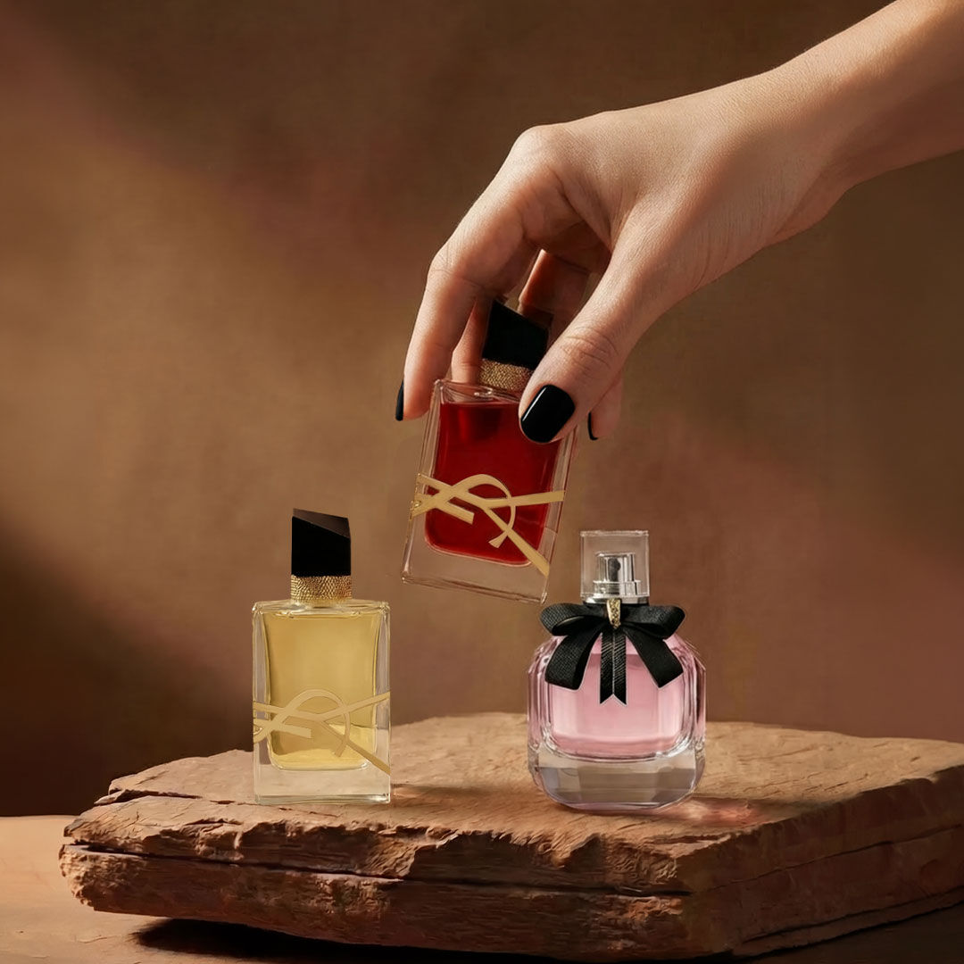 A hand holds a red YSL perfume bottle next to two other fragrances on a stone base.