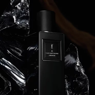 A sleek, rectangular black perfume bottle labeled "6 PLACE SAINT SULPICE LABDANUM" by YSL, set amongst striking pieces of shiny black marbled stone.
