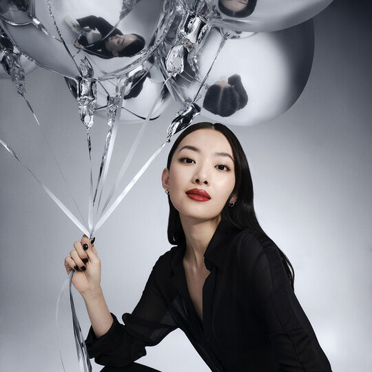 A woman with long dark hair, wearing a black shirt, holds silver balloons and looks directly at the camera. She wears red lipstick.