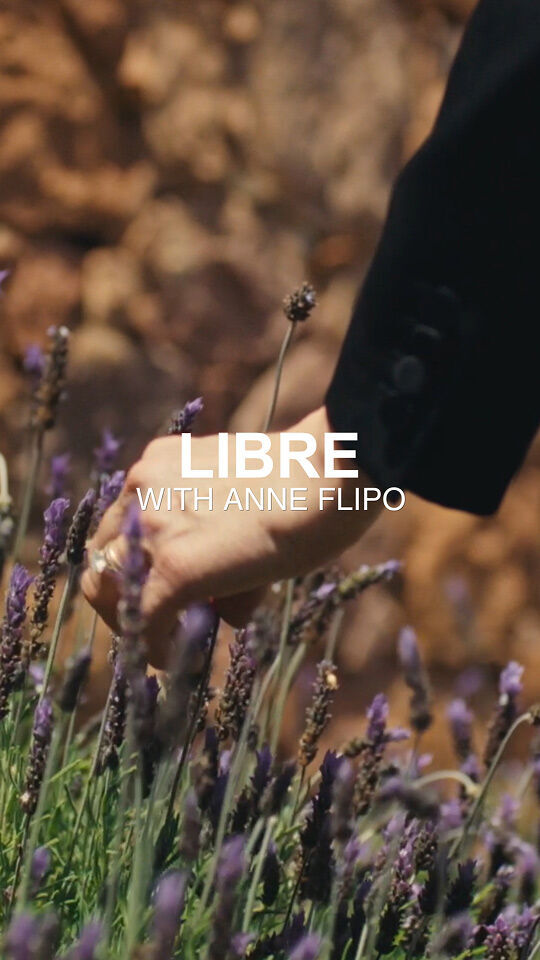 A hand, possibly belonging to Anne Flipo, reaches down towards a field of purple lavender flowers. The hand wears a ring and is partially obscured by the text overlay "LIBRE WITH ANNE FLIPO" in white. In the background, there is a blurry rock wall.