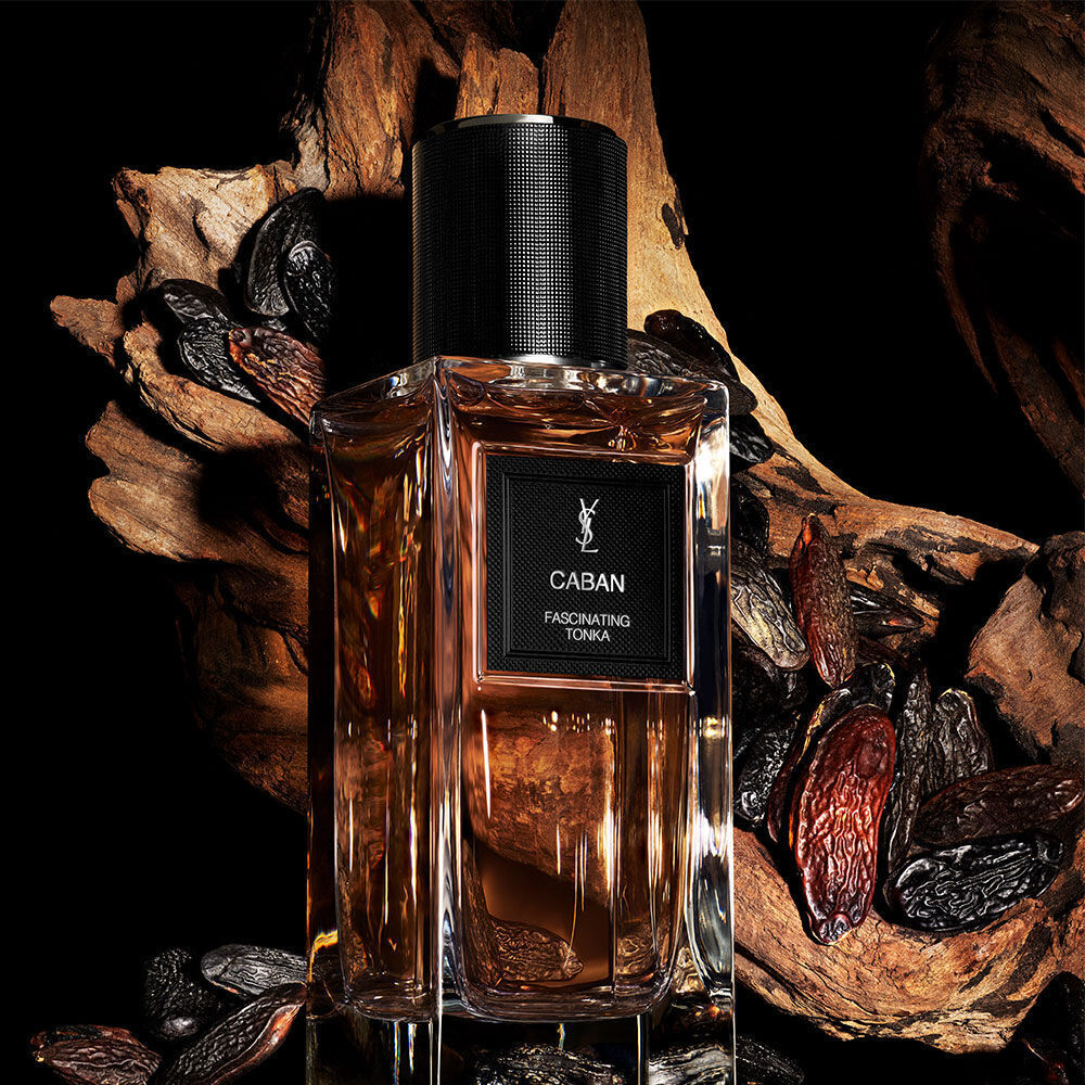 An amber-toned perfume bottle labeled "CABAN FASCINATING TONKA" by YSL, surrounded by tonka beans and a gnarled piece of driftwood.