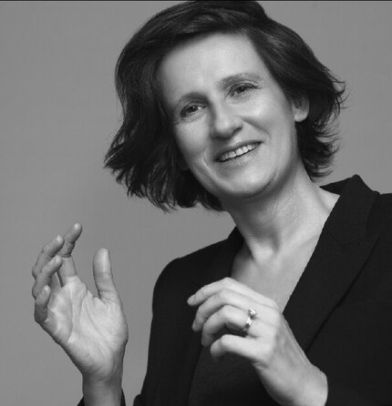 A woman with short, wavy hair is smiling and gesturing with her hands, wearing a black blazer against a neutral background.