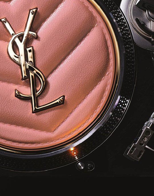 Close-up of a pink, quilted Yves Saint Laurent compact or accessory featuring a gold YSL logo at its center. The item is set against the metallic textured edge of a record player, blending luxury beauty with a music-inspired aesthetic.